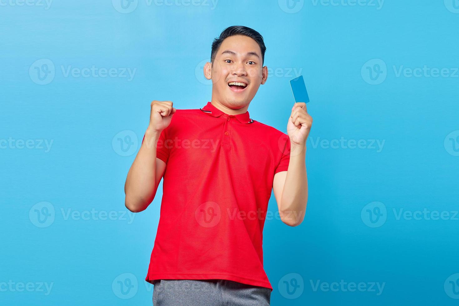 Portrait of excited young asian man holding a blank card and raising a fist in victory gesture isolated on blue background photo