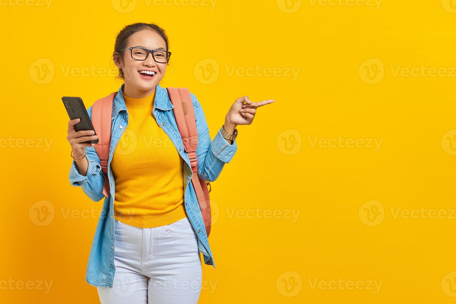 Portrait of  cheerful young Asian woman student in casual clothes with backpack holding mobile phone and pointing at copy space with fingers isolated on yellow background photo