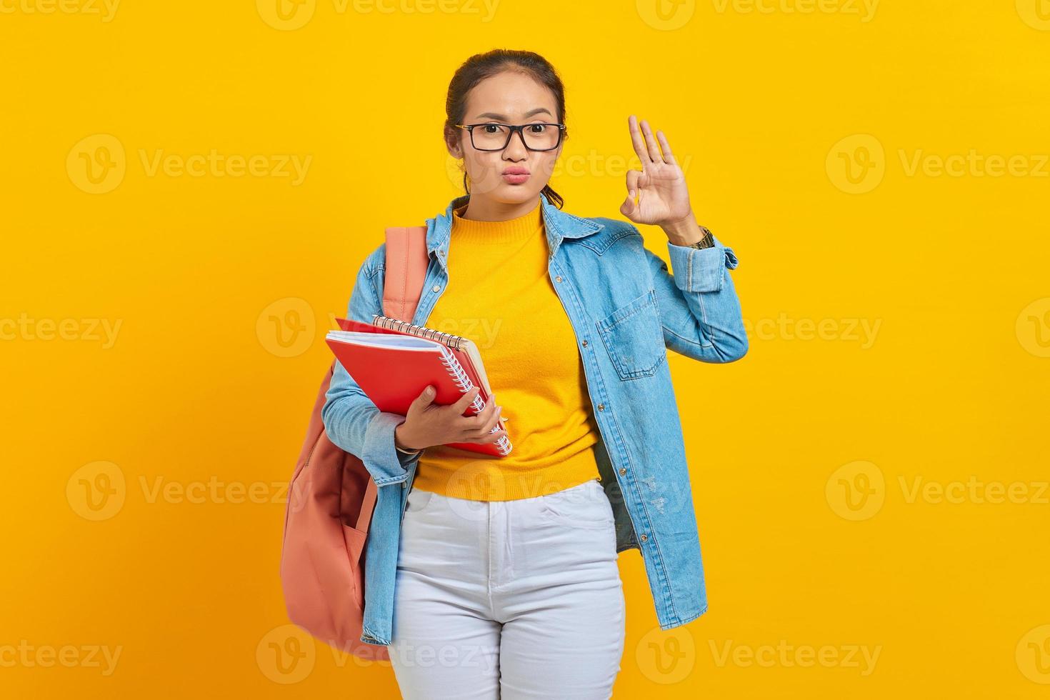 Funny young woman student in denim clothes with backpack, holding notebook and showing okay gesture isolated on yellow background. Education in high school university college concept photo