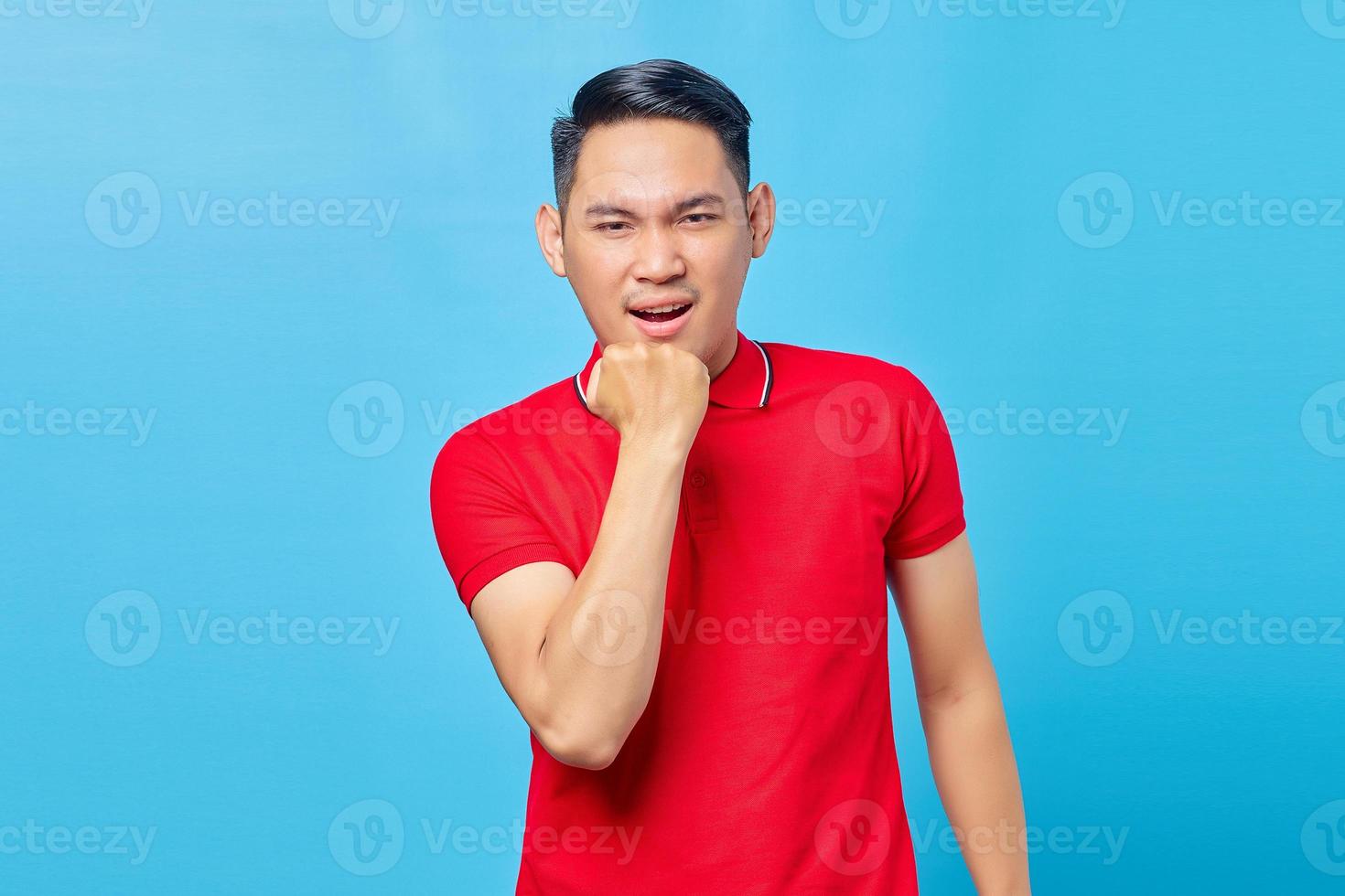 retrato de un joven asiático enojado y apuesto que gesticula y mantiene la boca abierta mientras está de pie con el puño aislado en el fondo azul foto