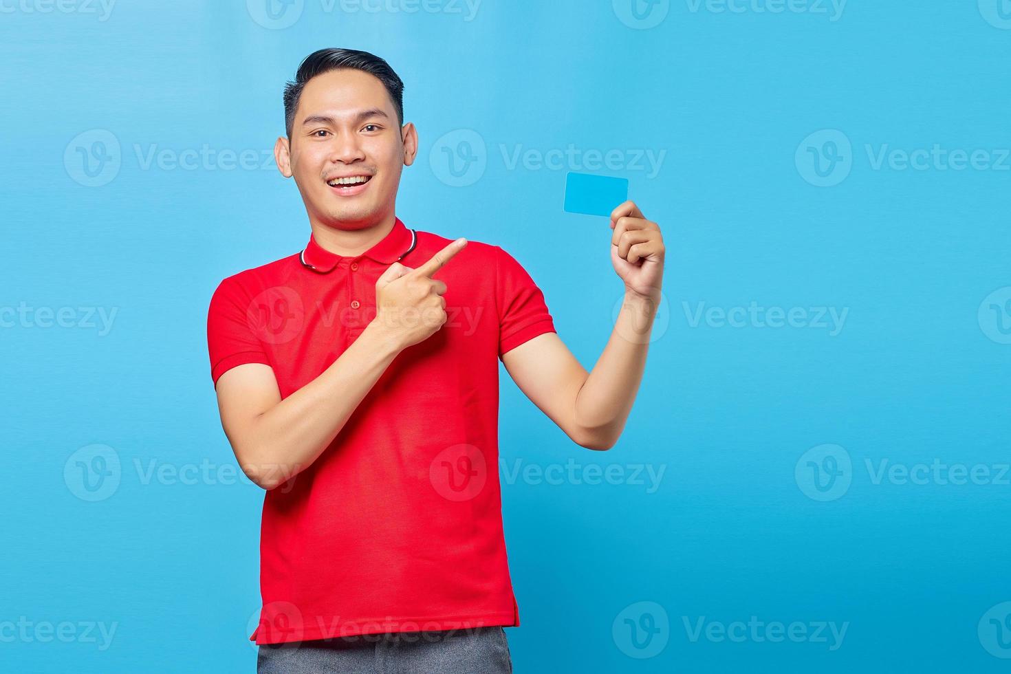 Portrait of cheerful young asian man pointing finger on credit card isolated on blue background photo