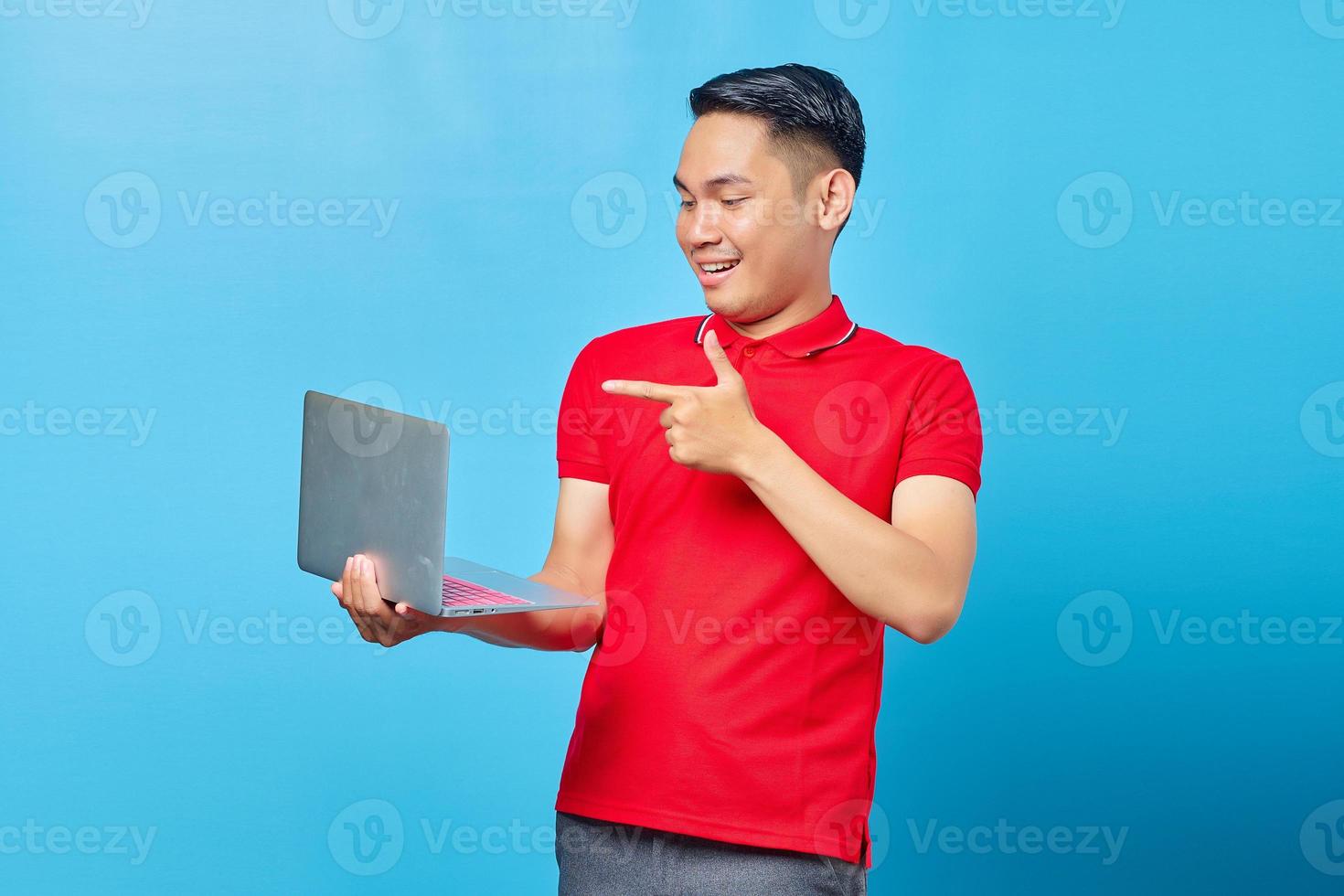 Portrait of cheerful Asian young man in red shirt pointing at laptop with finger isolated on blue background photo