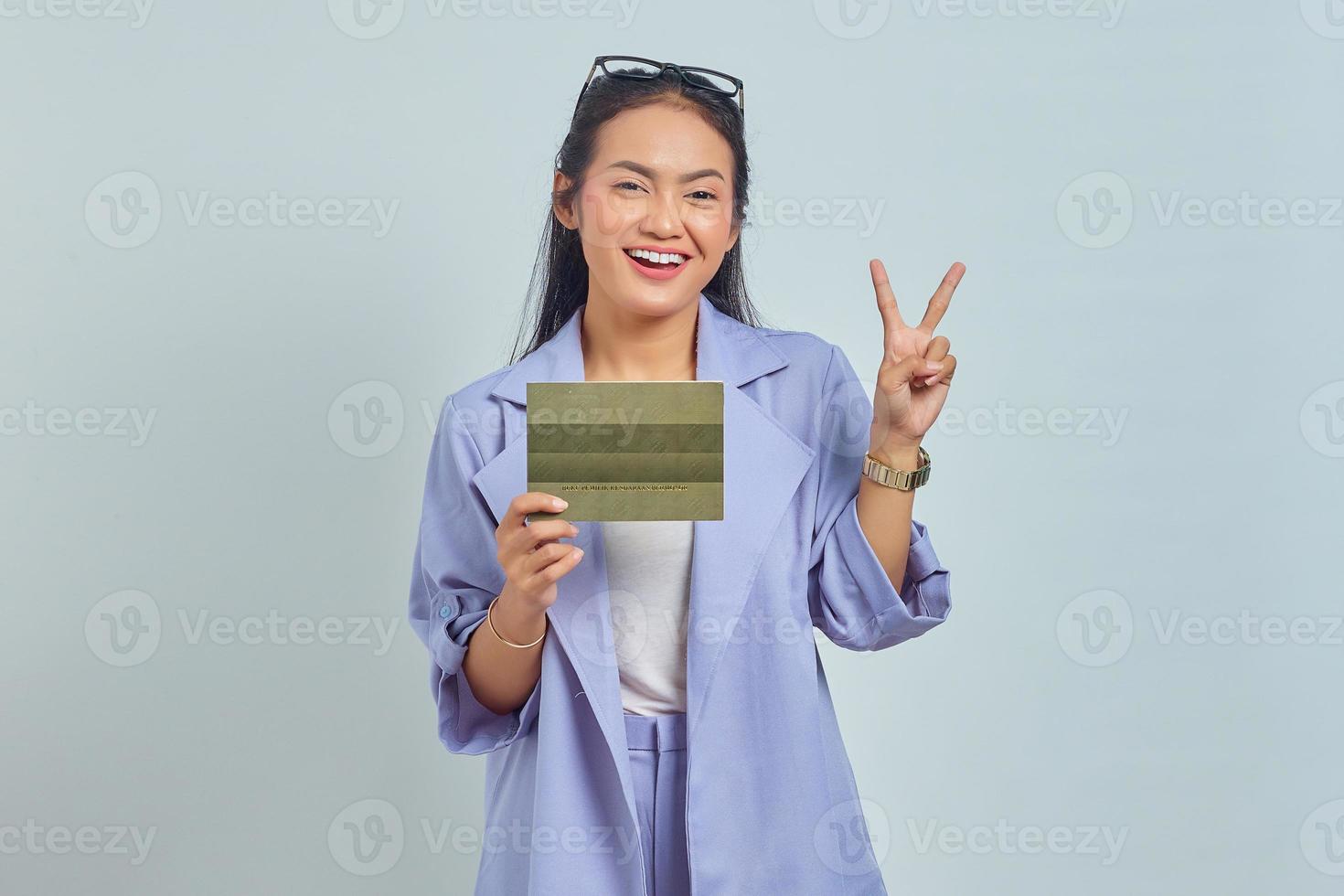 retrato de una alegre joven asiática sosteniendo un libro de vehículos y mostrando signos de paz con los dedos aislados en fondo blanco foto