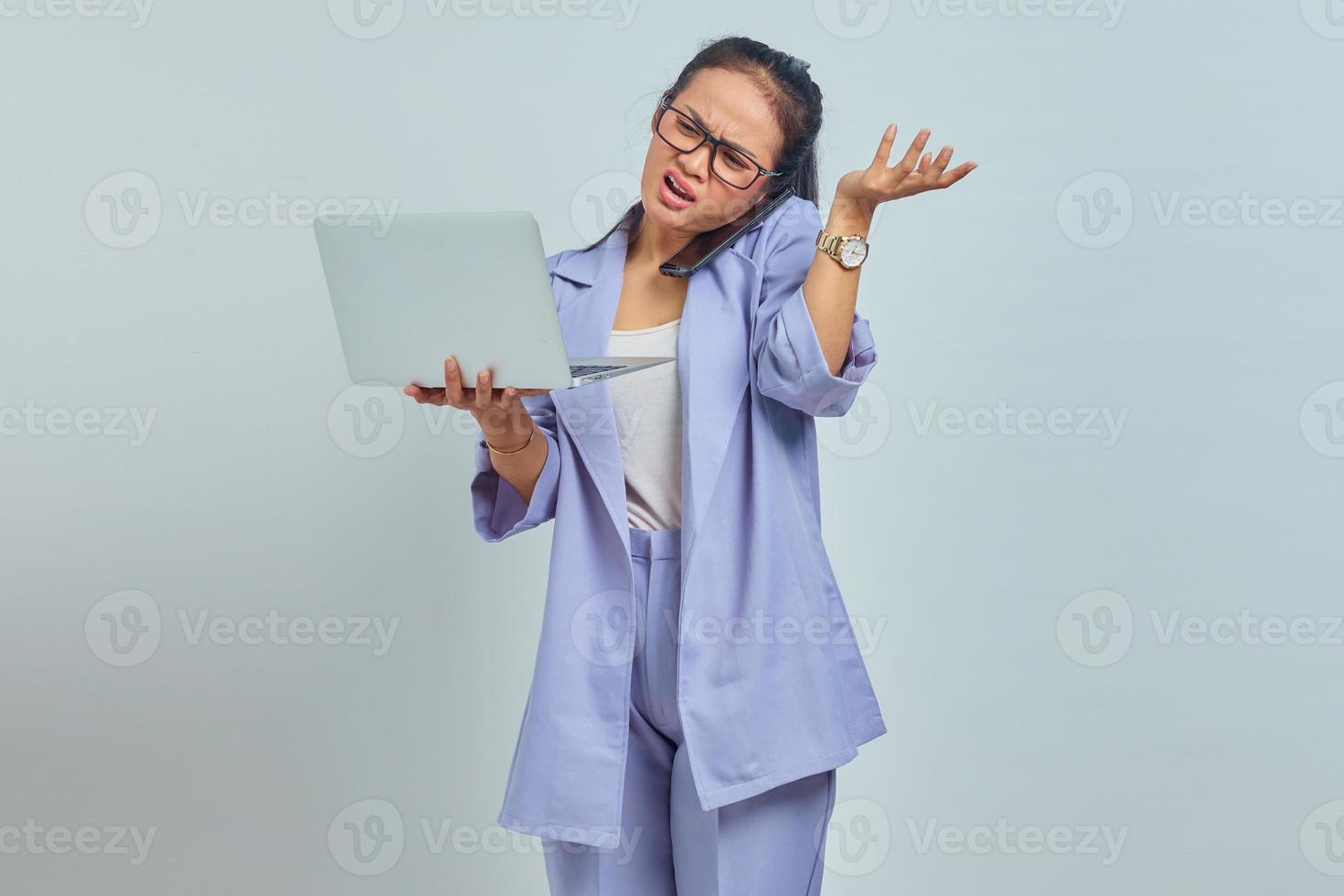Portrait of angry young Asian woman talking on mobile phone and holding laptop, pointing to copy space with palms isolated on white background photo