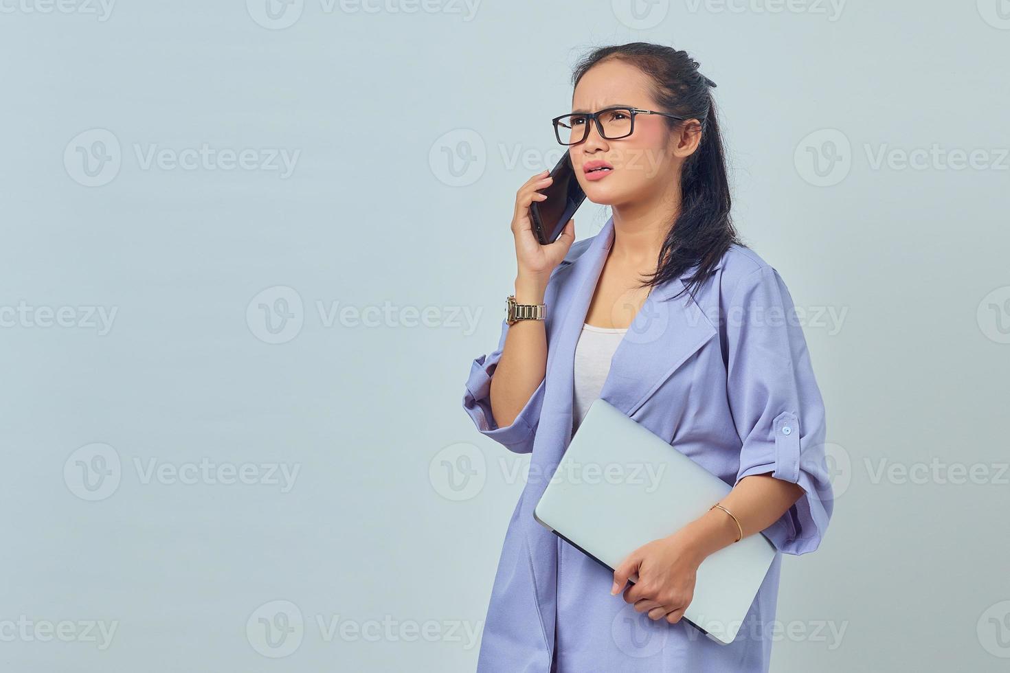 Portrait of confused young Asian woman talking on mobile phone and holding laptop isolated on white background photo