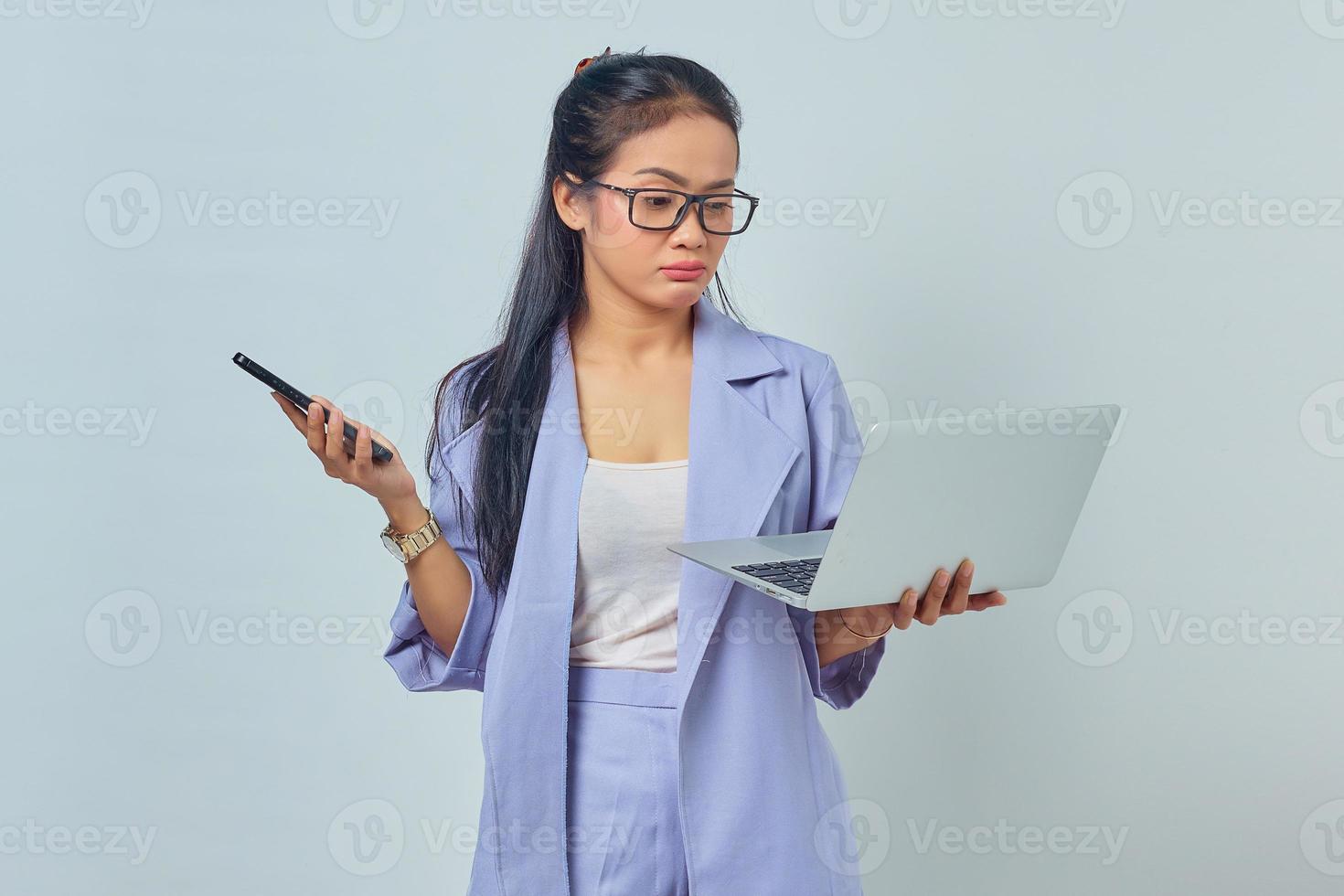 Portrait of young Asian woman standing using laptop and holding mobile phone with pensive expression isolated on white background photo