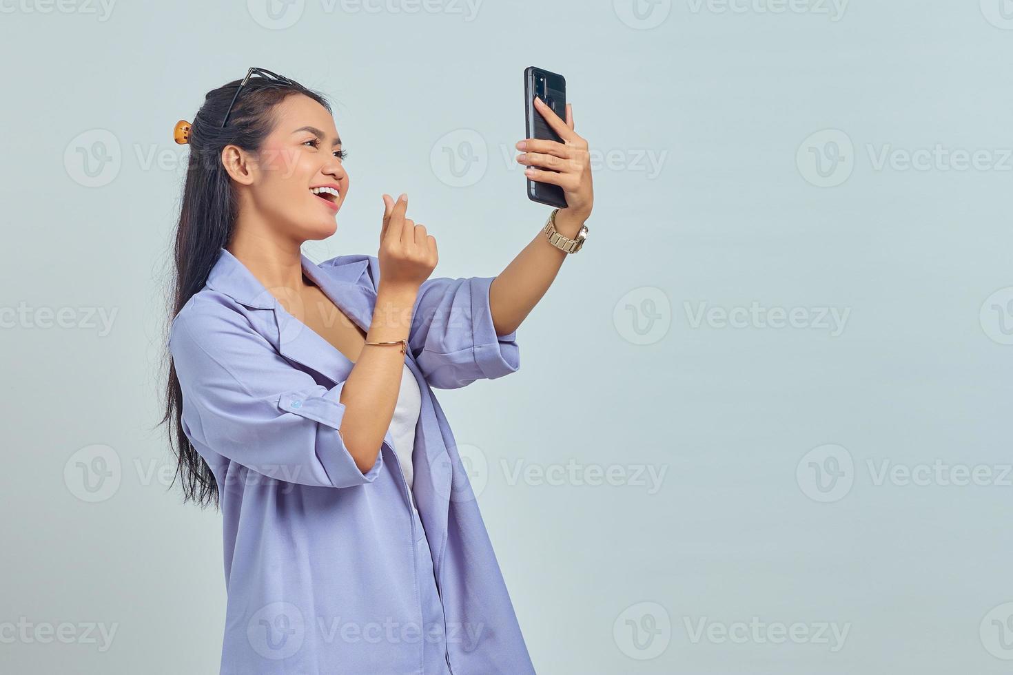 Portrait of cheerful young Asian woman using mobile phone taking selfie and showing heart sign with two fingers crossed isolated on white background photo