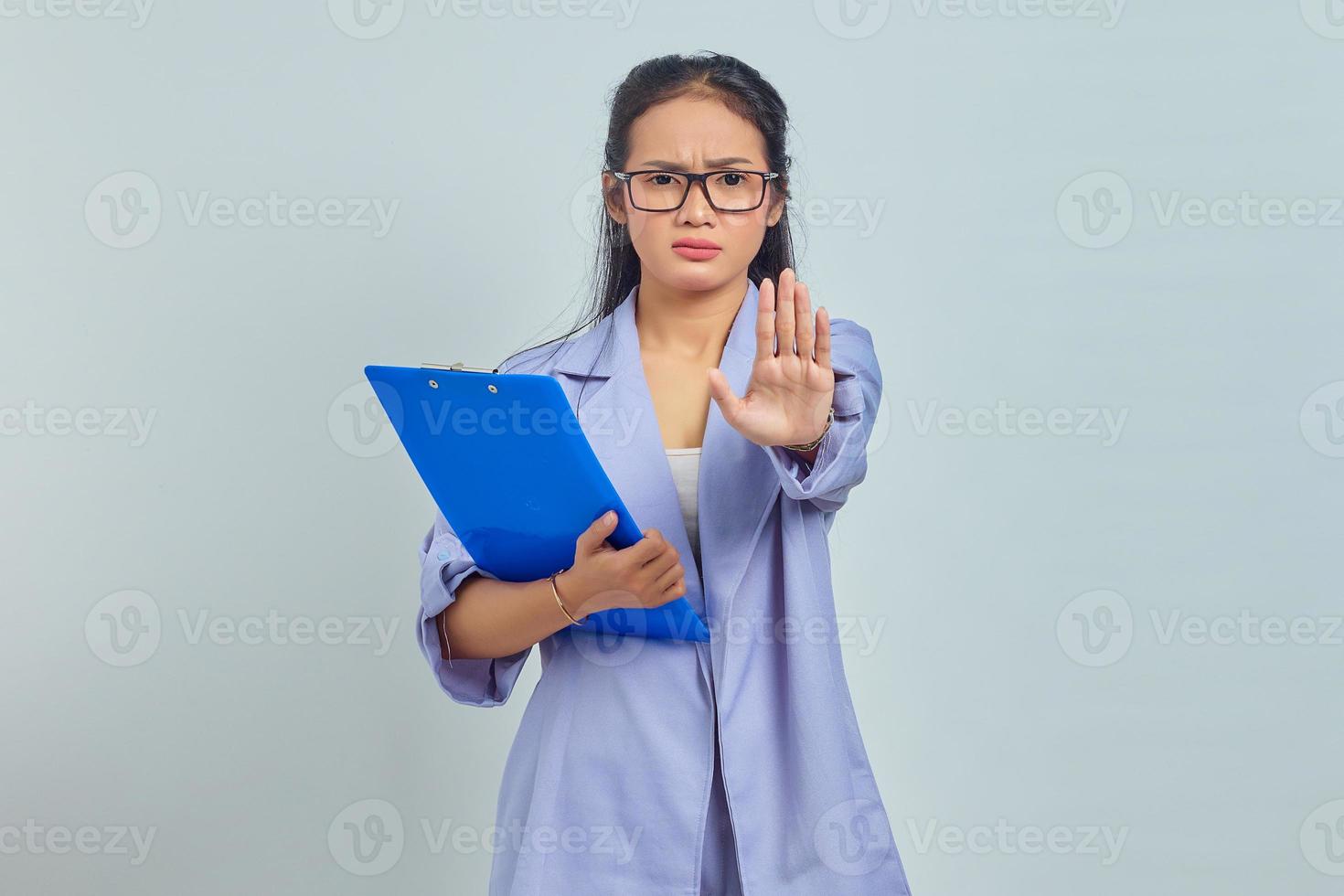 Portrait of angry young Asian woman holding folder and says no shows prohibition gesture refuses something isolated on purple background photo