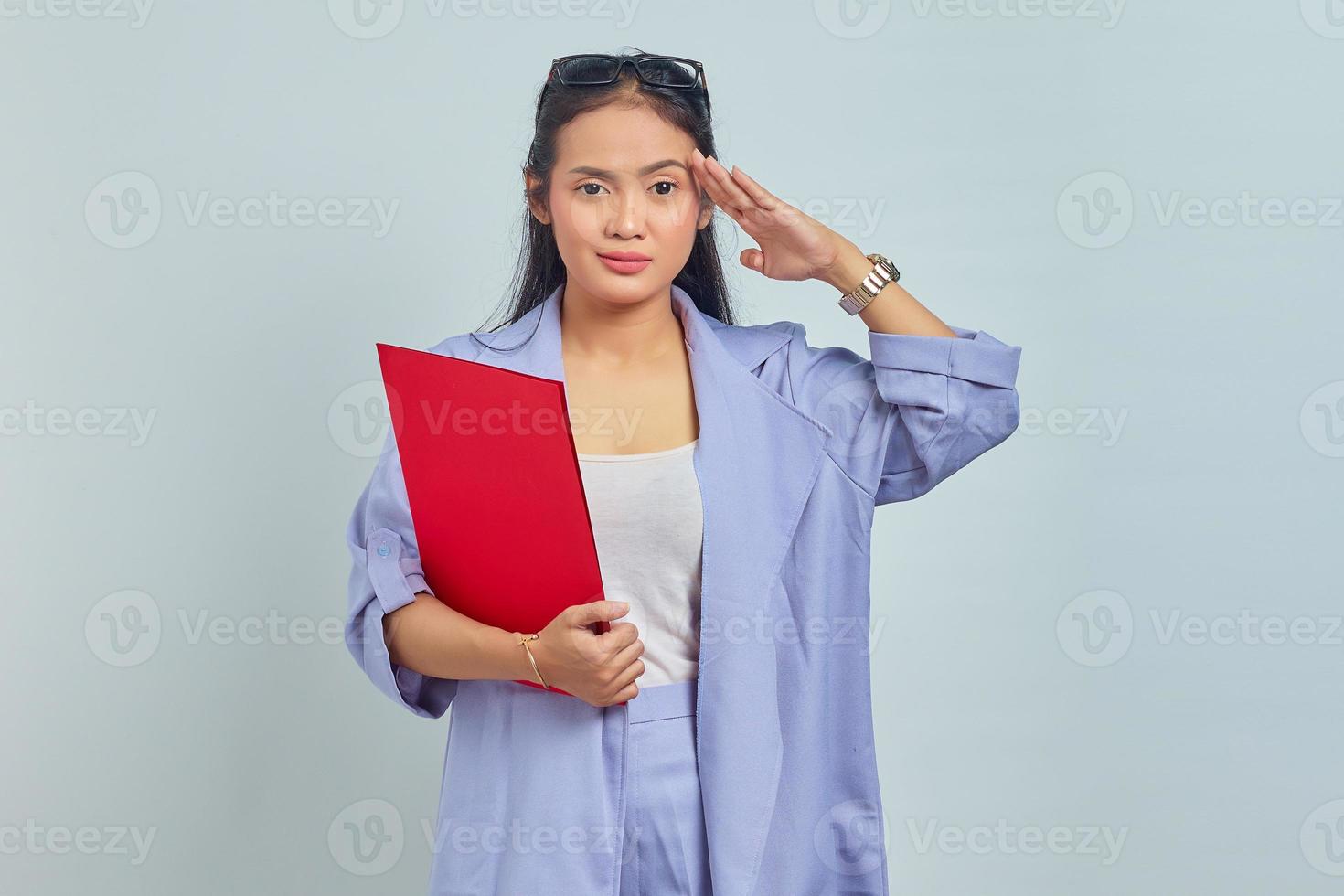 retrato de una joven asiática sonriente sosteniendo una carpeta de documentos y haciendo un gesto de respeto a un superior, lista para hacer trabajo de oficina aislada en un fondo morado foto