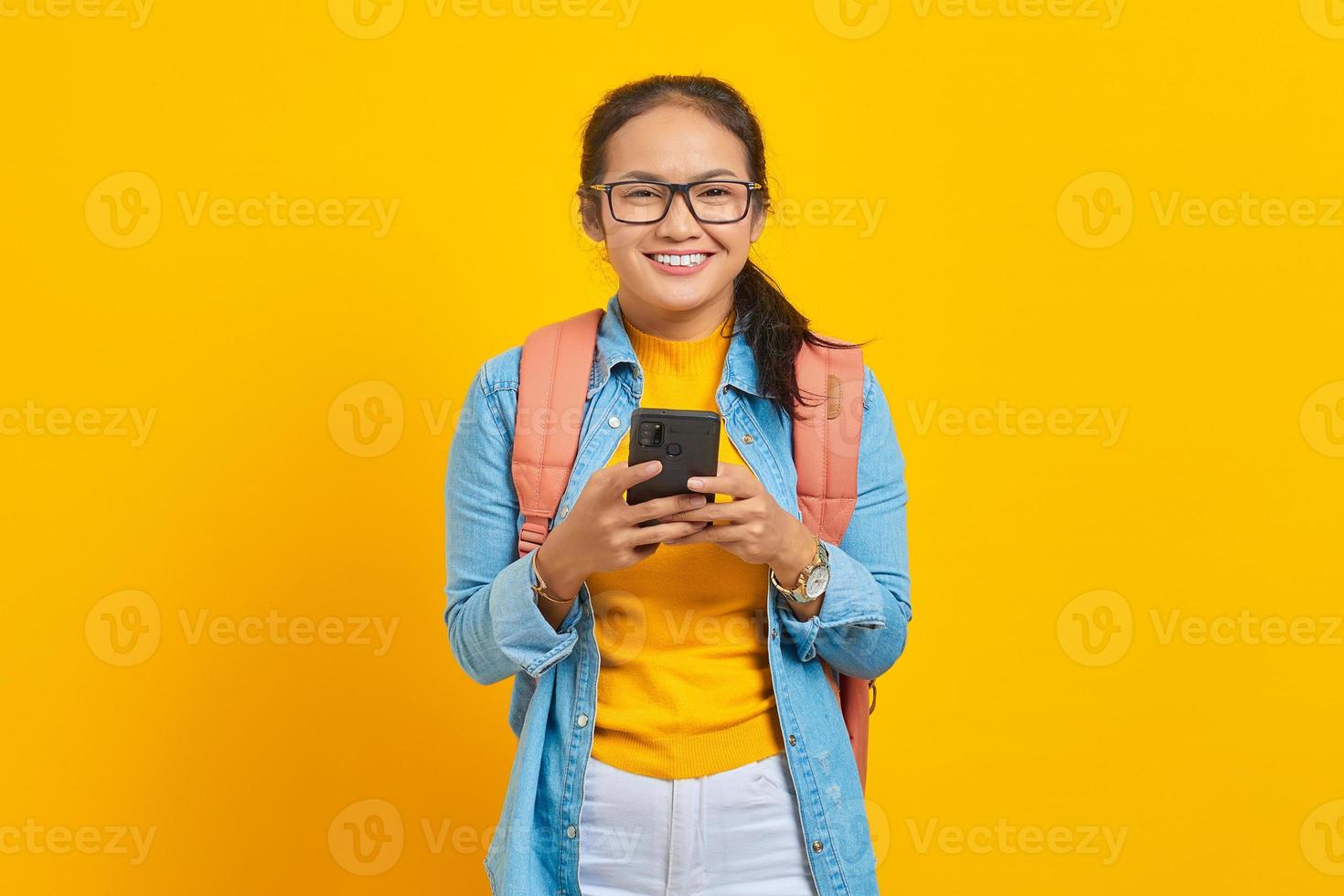 Portrait of cheerful young Asian woman student in casual clothes with backpack using mobile phone and looking at camera isolated on yellow background. Education in college university concept photo