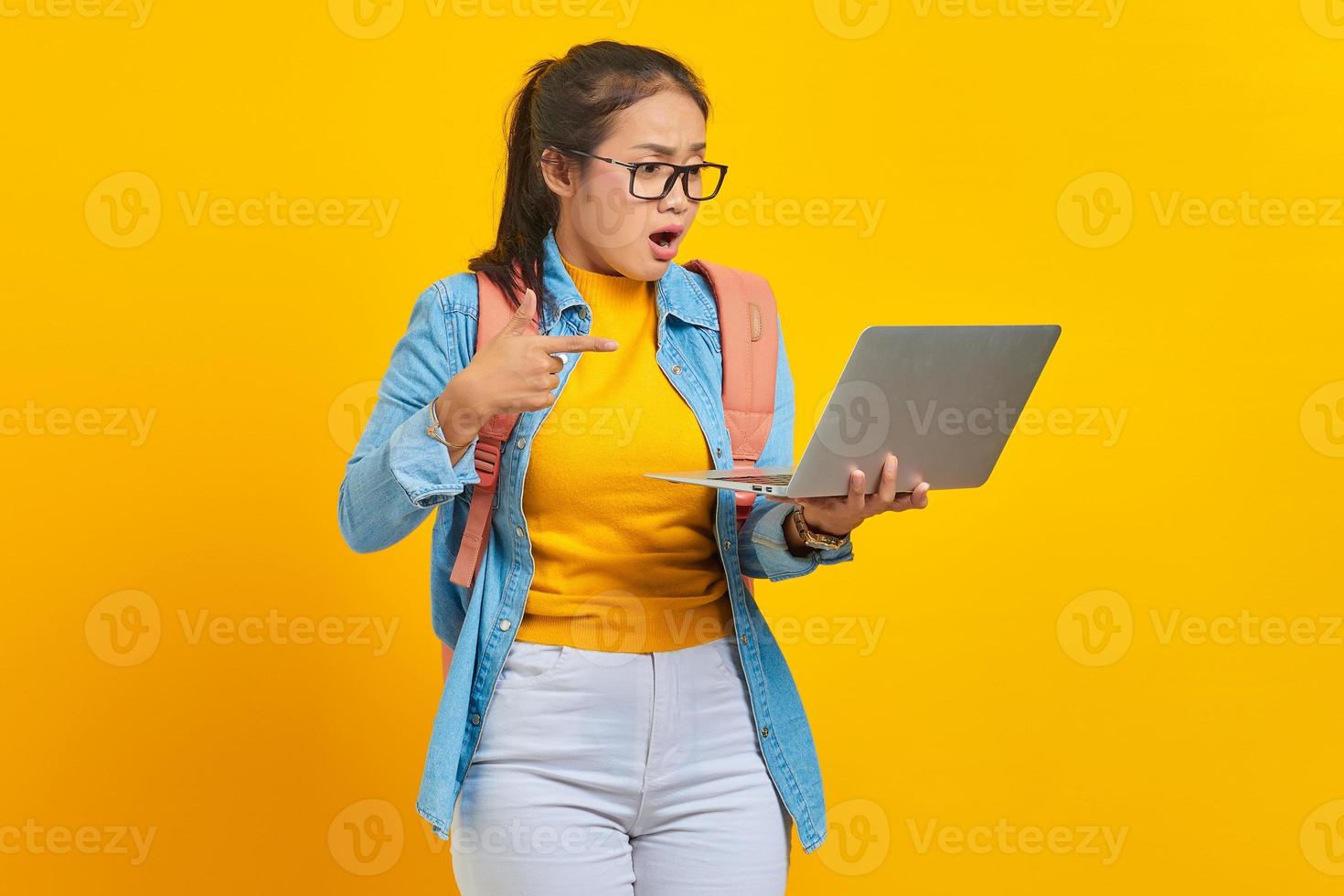 Portrait of surprised young Asian woman student in casual clothes with backpack pointing fingers at laptop isolated on yellow background. Education in university college concept photo