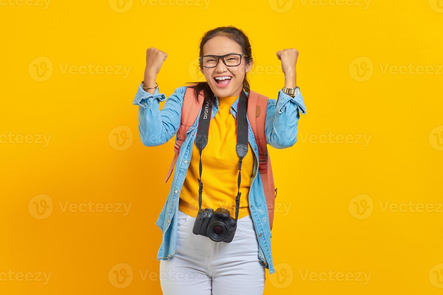 Portrait of excited young traveler Asian woman with backpack and camera in denim clothes while celebrate luck isolated on yellow background. Passenger traveling on weekends. Air flight journey concept photo