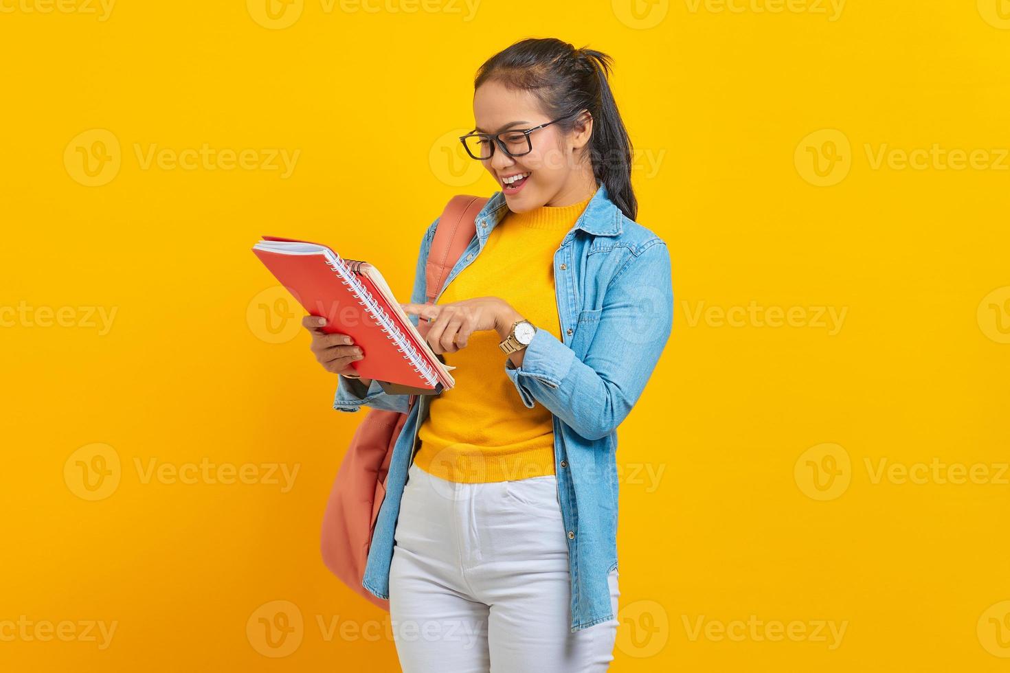 Smiling woman student in denim clothes with backpack,  reading in the work notebook isolated on yellow background. Education in high school university college concept photo