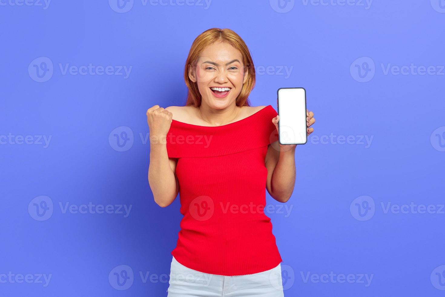 retrato de una joven asiática alegre que muestra un teléfono móvil con pantalla en blanco y hace un gesto de éxito aislado en un fondo morado foto