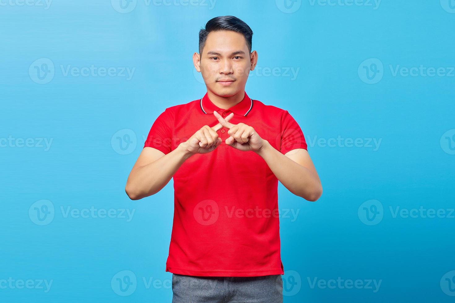 Portrait of serious handsome young man in red shirt crossing fingers and looking at camera with showing rejection attitude on blue background photo