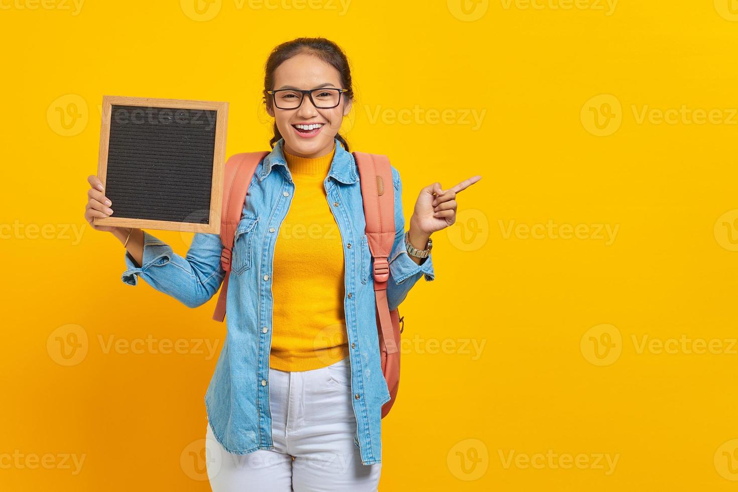 Portrait of smiling young Asian woman student in casual clothes with backpack holding blank chalkboard and pointing at copy space with finger isolated on yellow background photo