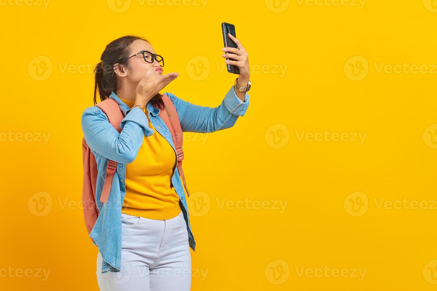 Portrait of  cheerful young Asian woman student in casual clothes with backpack having video call and hold hand palms send air kiss to boyfriend at smartphone camera isolated on yellow background photo
