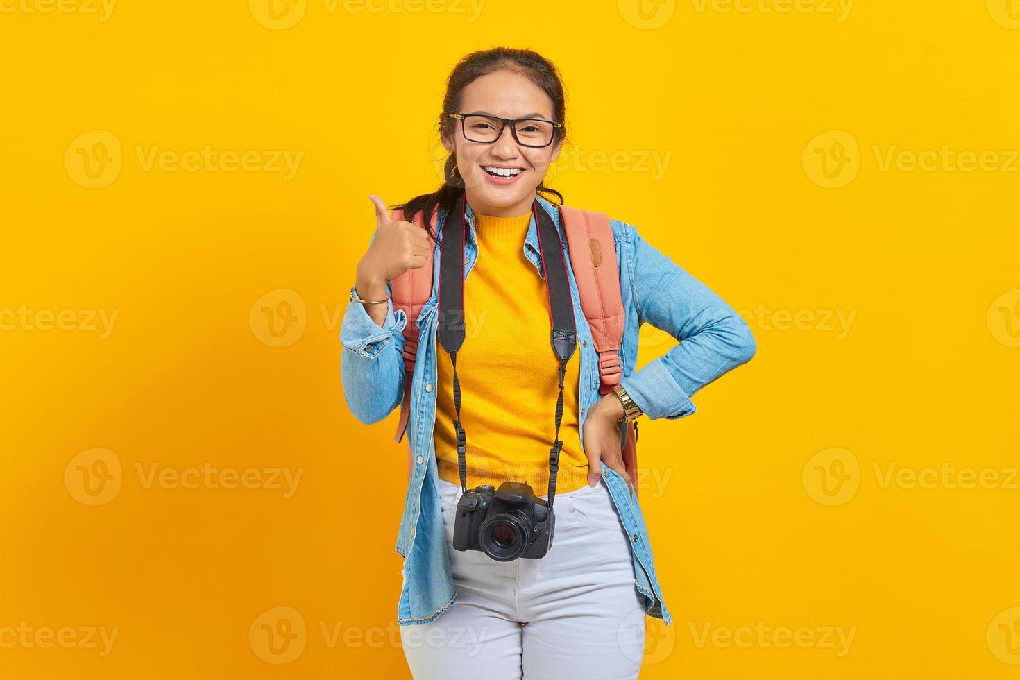 retrato de una alegre joven viajera asiática con mochila y cámara vestida de denim mientras muestra un gesto con el pulgar hacia arriba aislado en un fondo amarillo. concepto de viaje de vuelo aéreo foto