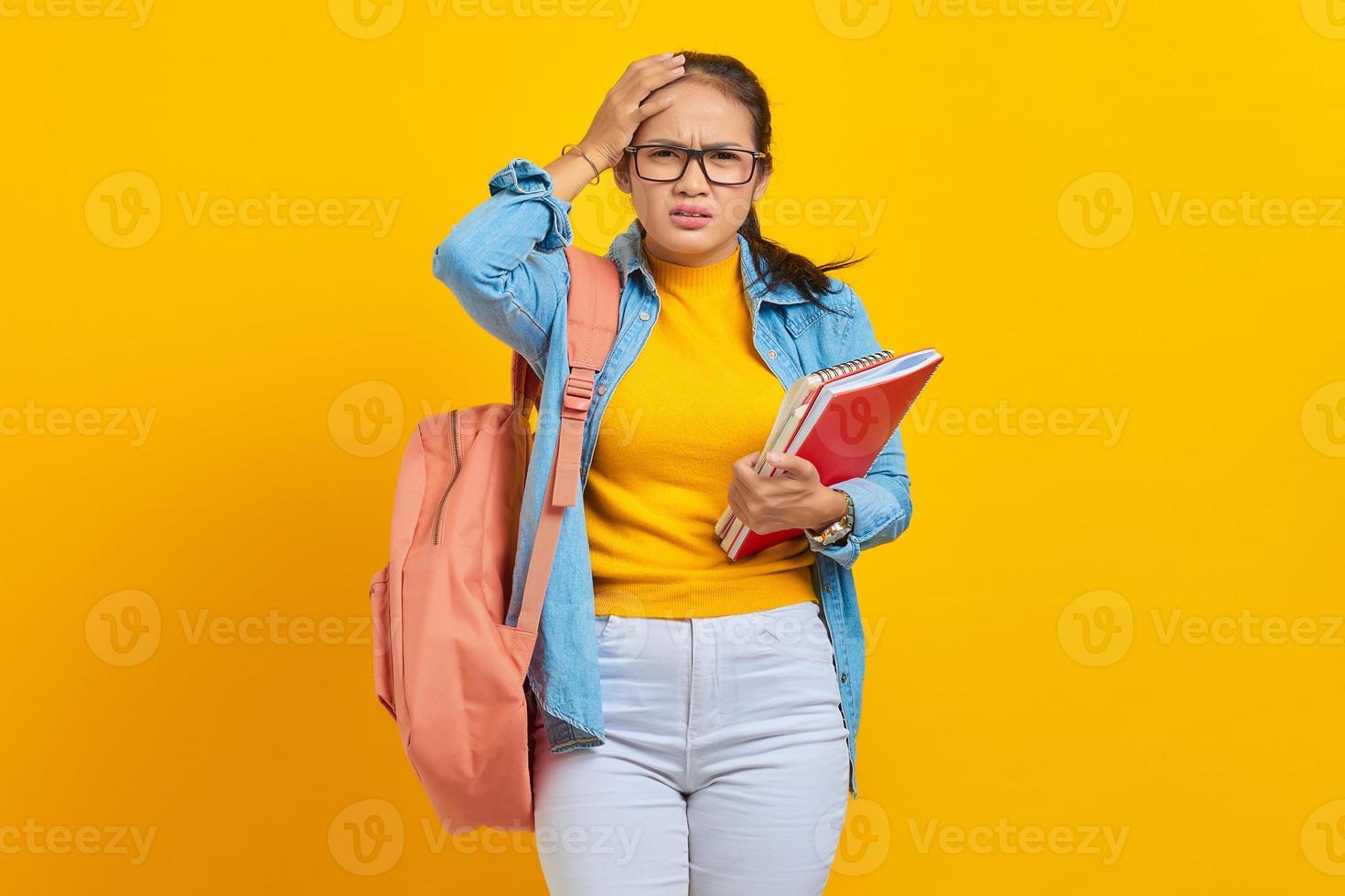 Beautiful young Asian college student in denim outfit with backpack holding notebook and looking dizzy with headache isolated on yellow background. Education in high school university college concept photo