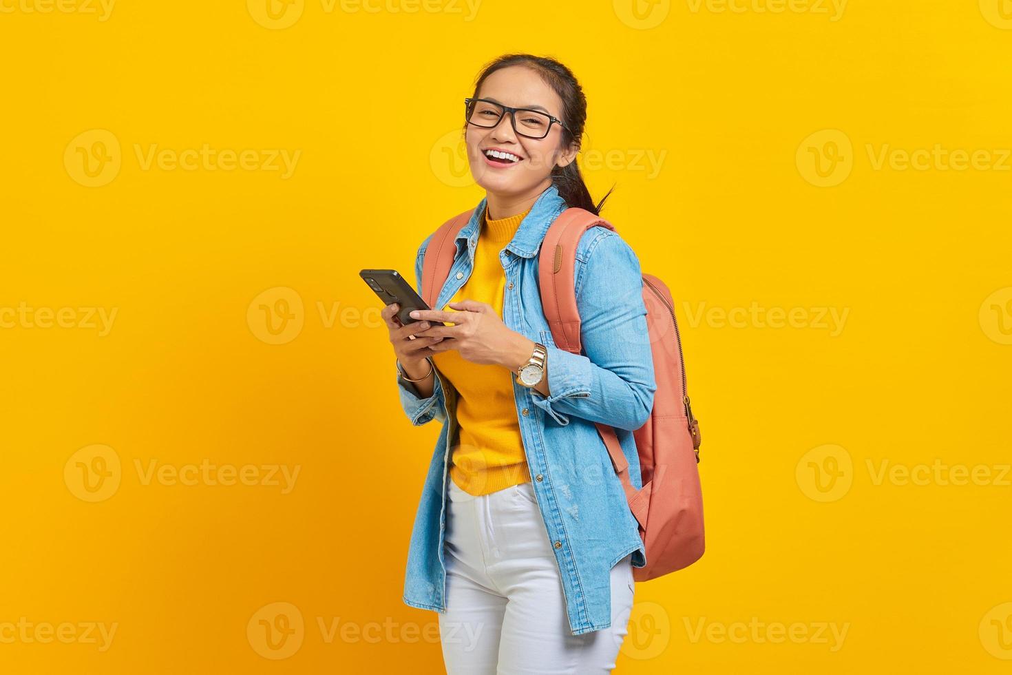 Portrait of cheerful young Asian woman student in casual clothes with backpack using mobile phone and looking at camera isolated on yellow background. Education in college university concept photo