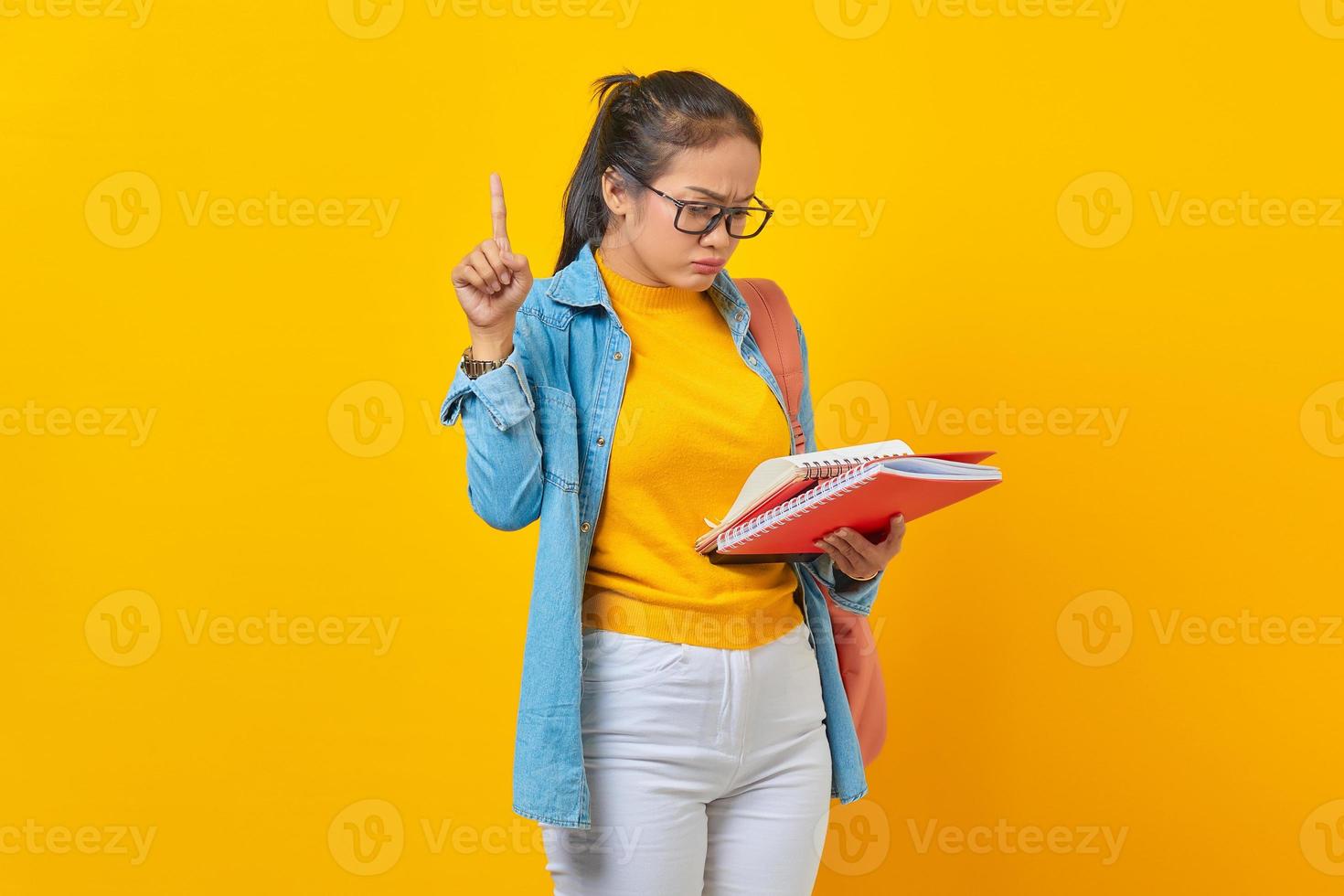 joven estudiante vestida de denim con mochila, sosteniendo un libro apuntando con el dedo índice hacia arriba con una gran idea nueva aislada en un fondo amarillo. educación en el concepto de colegio universitario de secundaria foto