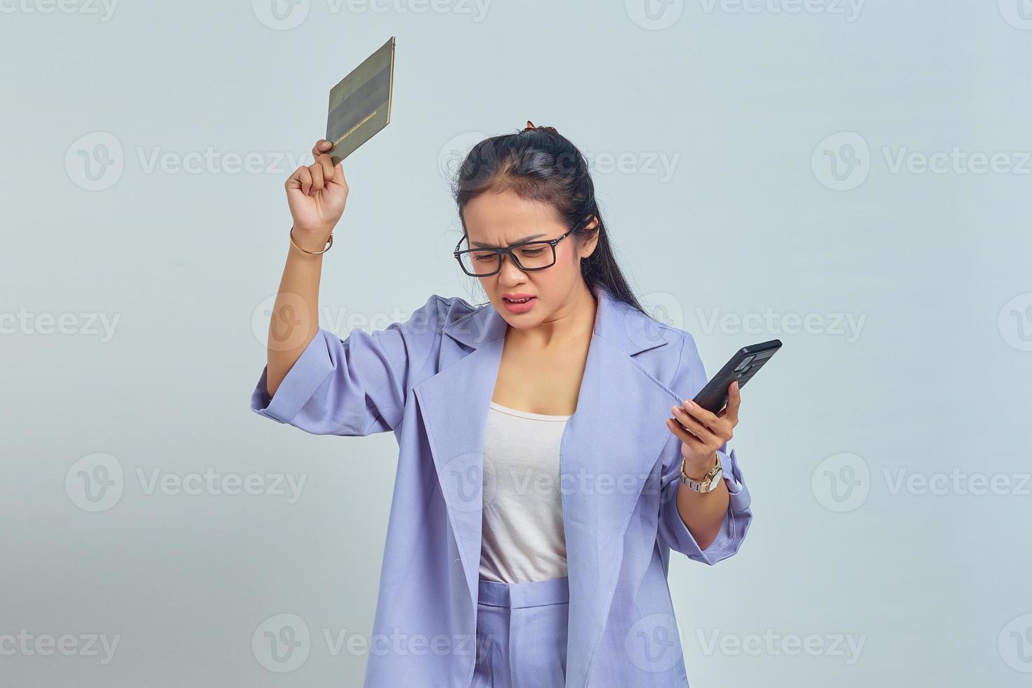 Portrait of angry young Asian woman with gesture of throwing away vehicle book and holding smartphone isolated on white background photo