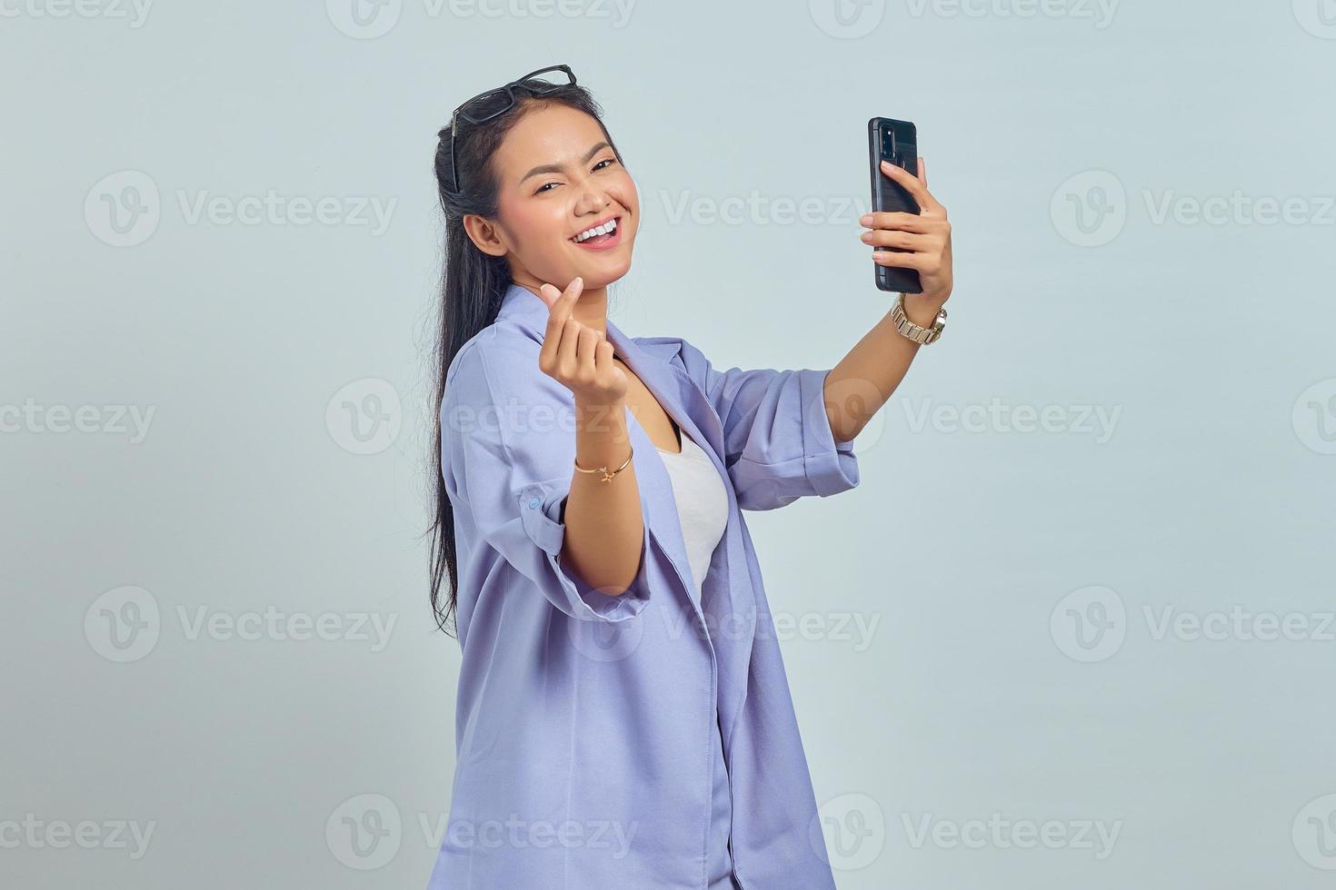 Portrait of cheerful young Asian woman using mobile phone taking selfie and showing heart sign with two fingers crossed isolated on white background photo