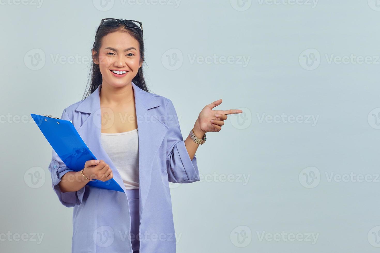 Portrait of smiling young Asian woman holding document folder and pointing finger at copy space isolated on purple background photo