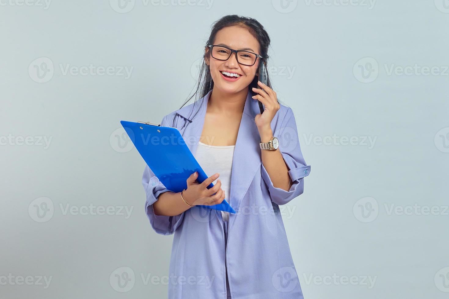 Portrait of cheerful young Asian woman laughing while talking on mobile phone with friends about something funny while holding document folder isolated on purple background photo