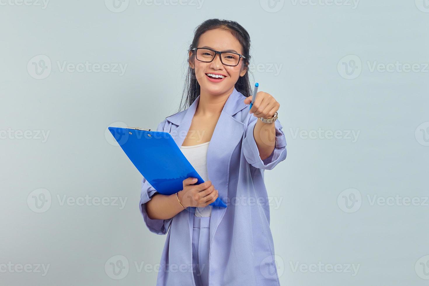 retrato de una hermosa joven mujer de negocios asiática con traje de pie sosteniendo una carpeta de documentos y señalando con un bolígrafo mirando a la cámara con cara sonriente aislada en un fondo morado foto