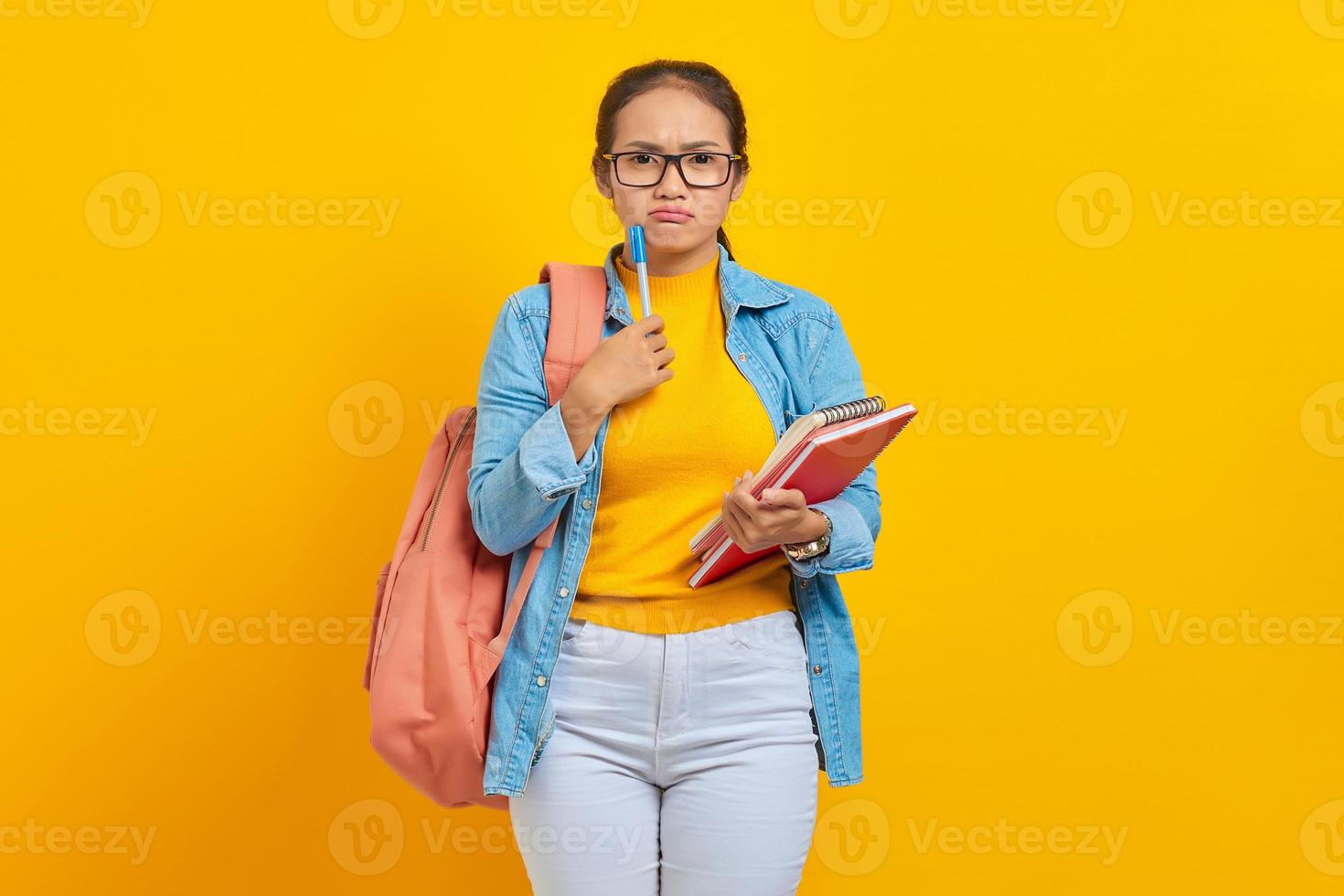 Portrait of serious young Asian woman student in casual clothes with backpack holding book and pen, thinking about question isolated on yellow background. Education in college university concept photo