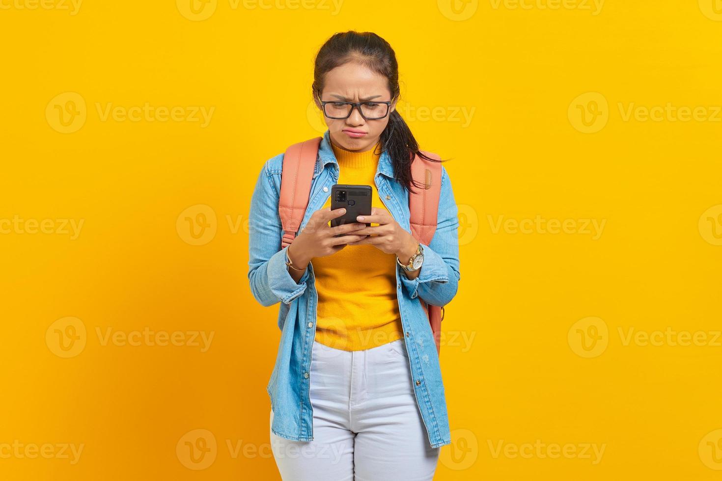 retrato de una joven estudiante asiática molesta vestida de forma informal con mochila usando un teléfono móvil, escribiendo mensajes sms aislados en un fondo amarillo. educación en concepto de universidad universitaria foto