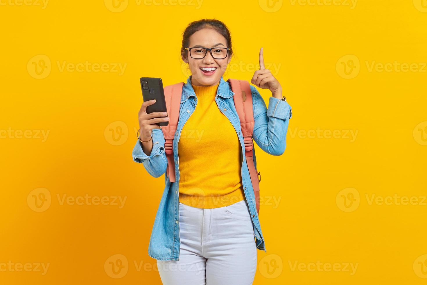 retrato de una alegre joven estudiante asiática vestida de forma informal con mochila sosteniendo un teléfono móvil y señalando el espacio con los dedos aislados en un fondo amarillo foto