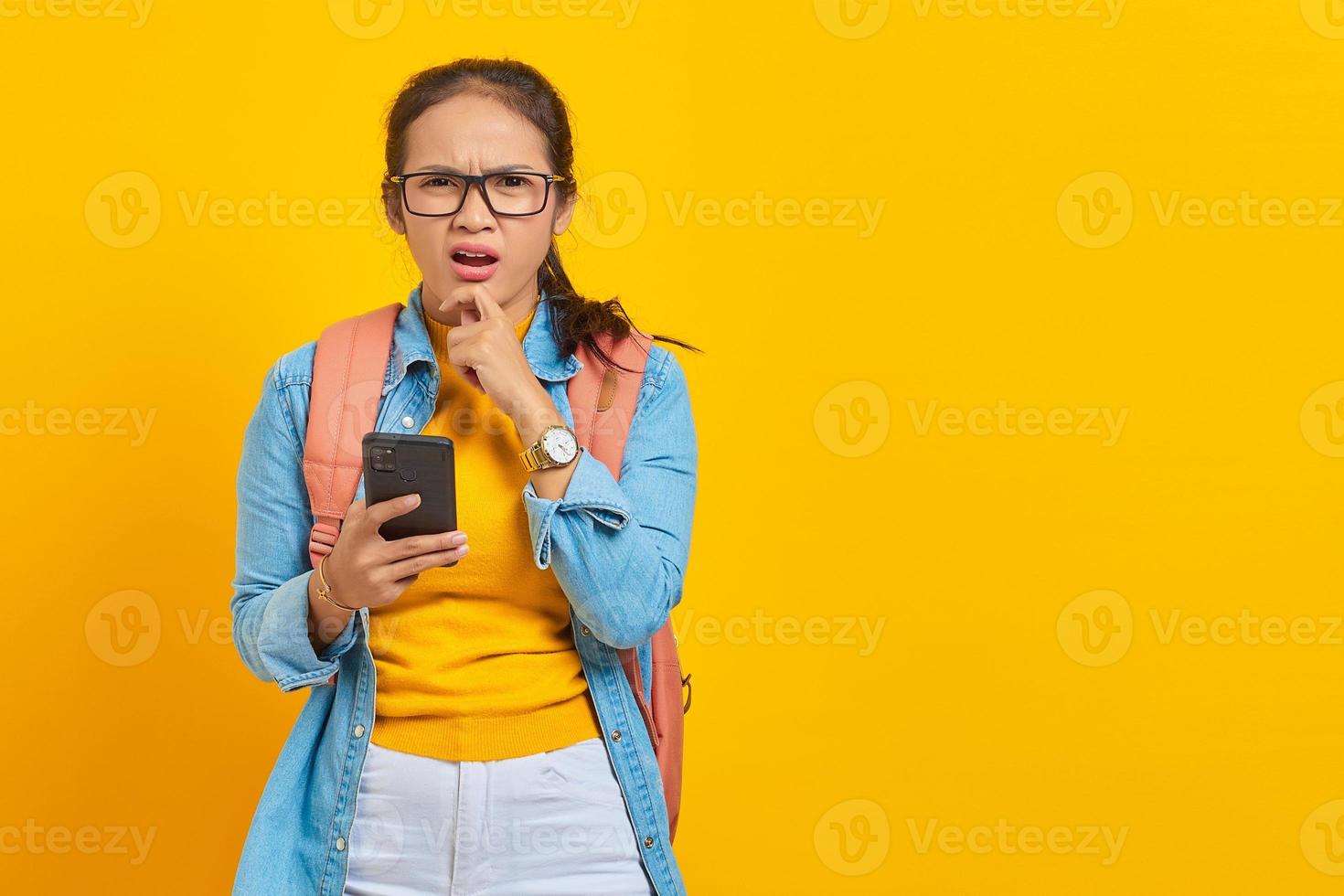 Portrait of pensive young Asian woman student in casual clothes with backpack using mobile phone and holding chin while thinking something isolated on yellow background photo