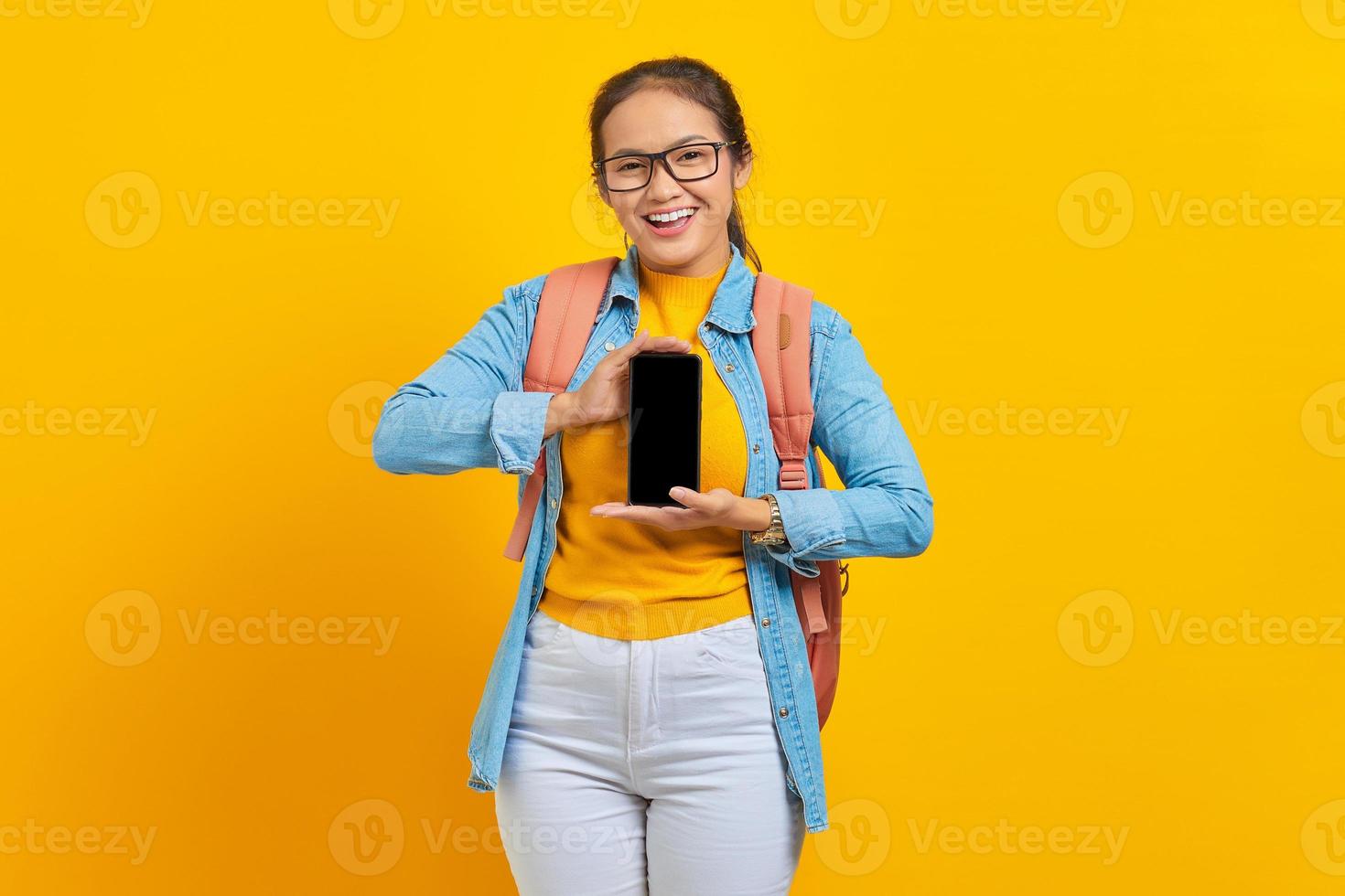 Portrait of cheerful young Asian woman student in casual clothes with backpack  showing blank screen mobile phone isolated on yellow background. Education in college university concept photo