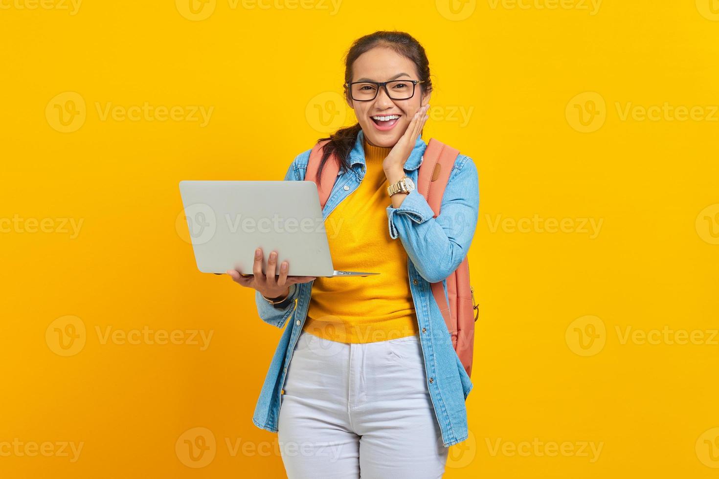 Portrait of excited young Asian woman student in casual clothes with backpack using laptop and  touching cheek with hands isolated on yellow background. Education in college university concept photo