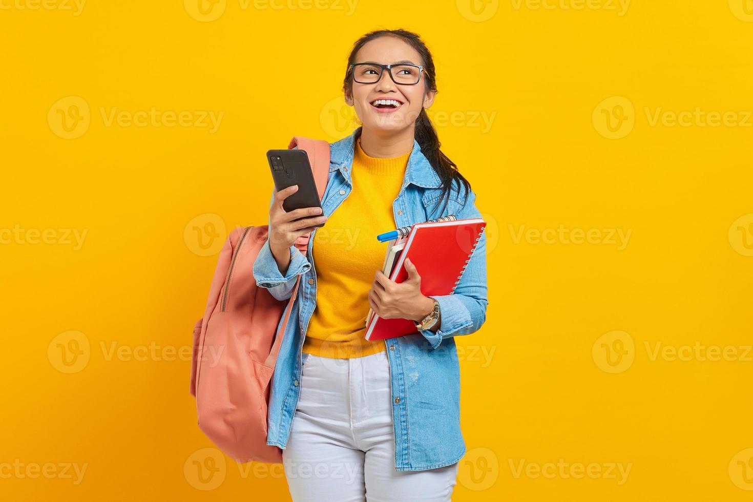 Portrait of smiling young Asian woman student in denim clothes, glasses with backpack holding mobile phone and books isolated on yellow background. Education in high school university college concept photo