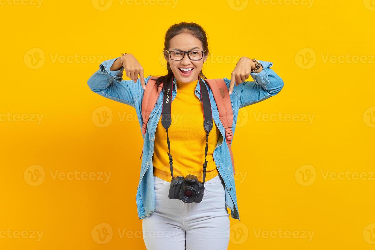 Portrait of cheerful young traveler Asian woman with backpack and camera in denim clothes while pointing at copy space with fingers isolated on yellow background. Air flight journey concept photo