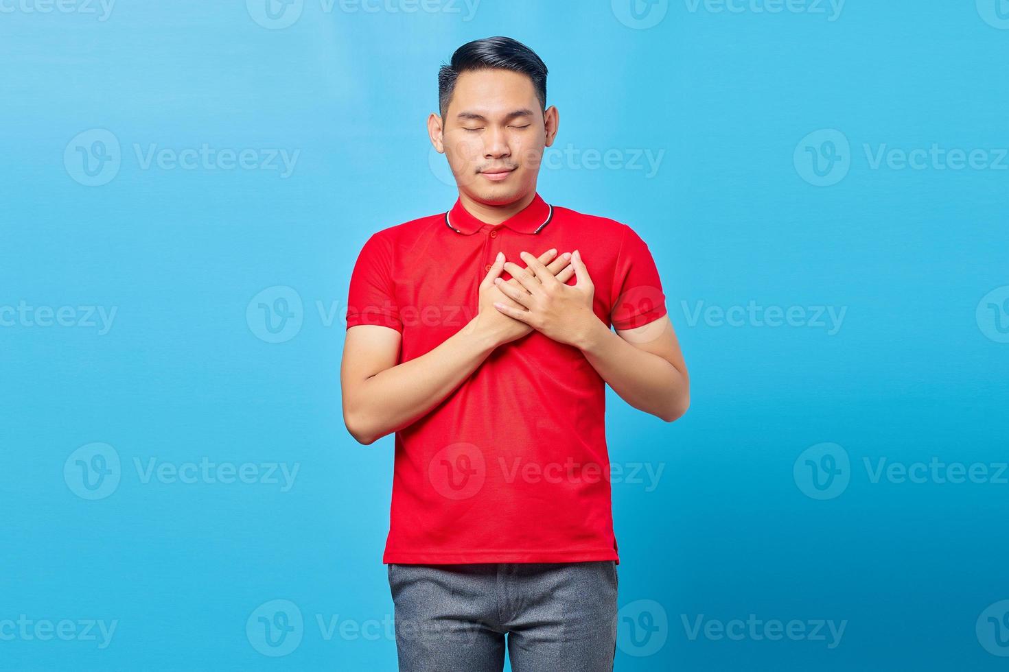 Portrait of handsome young Asian man in red shirt pressing hands to chest with closed eyes and grateful gesture on face. Health concept. photo