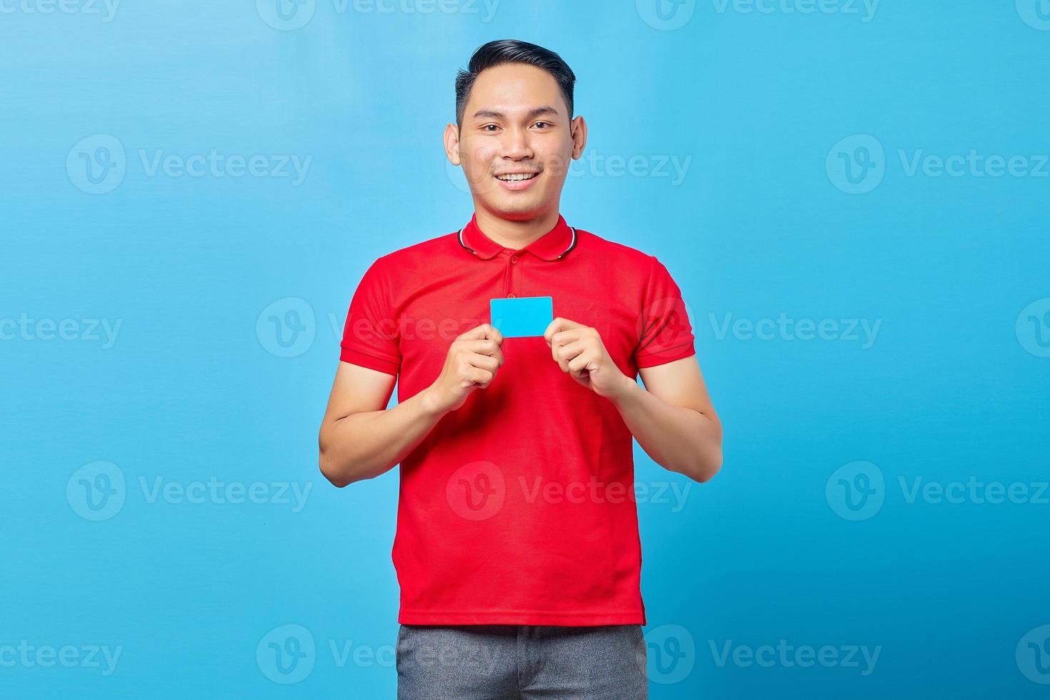 retrato de un joven asiático emocionado que muestra una tarjeta de crédito en blanco y mira una cámara aislada de fondo azul foto