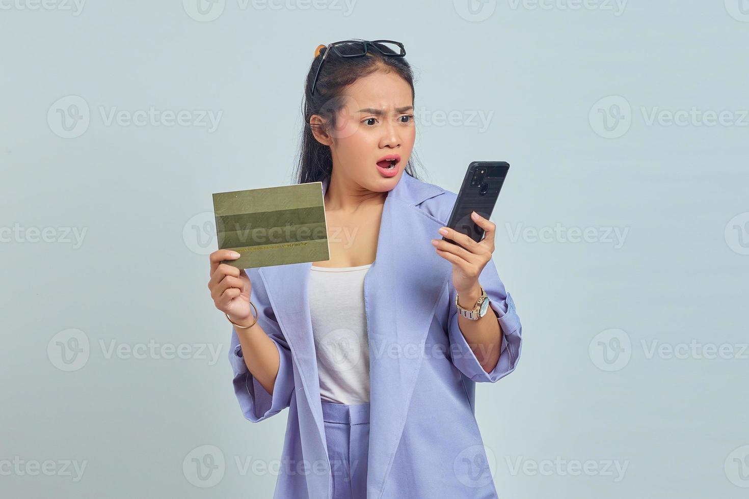 Portrait of surprised young Asian woman showing vehicle book and holding smartphone isolated on white background photo
