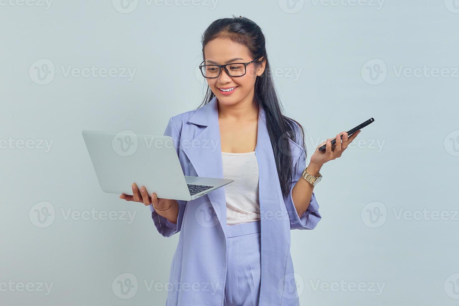Portrait of young Asian woman standing using laptop and holding mobile phone with cheerful expression isolated on white background photo