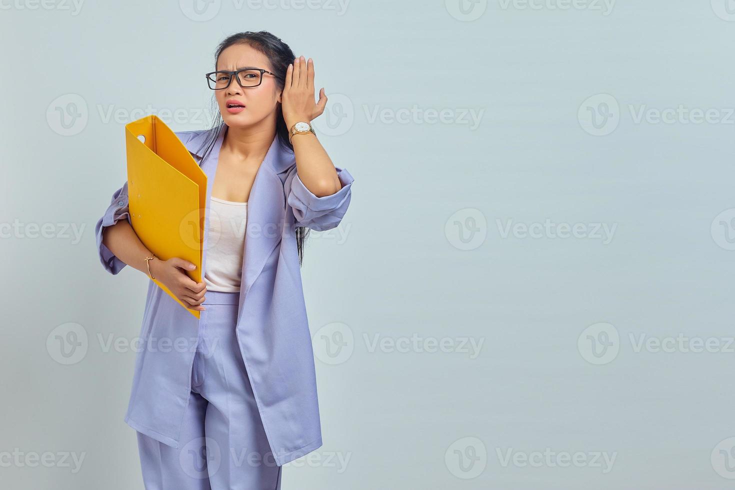 Portrait of serious young Asian business woman keeps hand near ear tries to overhear private conversation listens gossips while holding yellow folder isolated on purple background photo