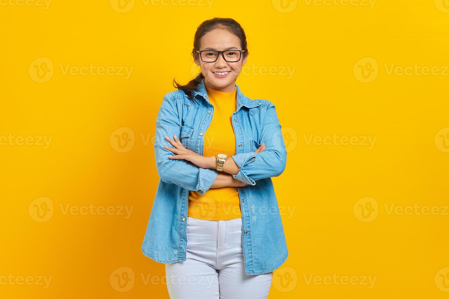 retrato de una joven estudiante asiática sonriente vestida de denim con los brazos cruzados en el pecho y luciendo confiada aislada en un fondo amarillo. educación en concepto de universidad universitaria foto