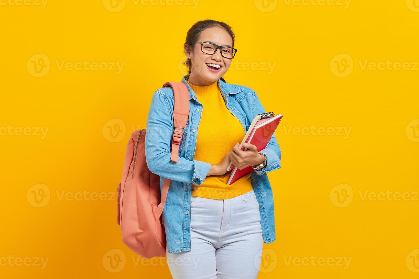 retrato de una alegre joven estudiante asiática vestida de forma informal con mochila sosteniendo un libro y mirando una cámara aislada de fondo amarillo foto