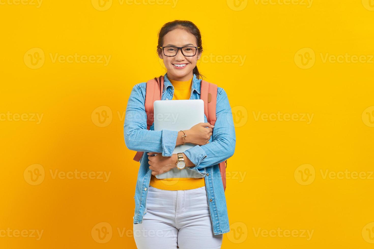 retrato de una joven estudiante asiática sonriente con ropa informal con mochila sosteniendo una laptop y mirando una cámara aislada en un fondo amarillo. educación en concepto de colegio universitario foto