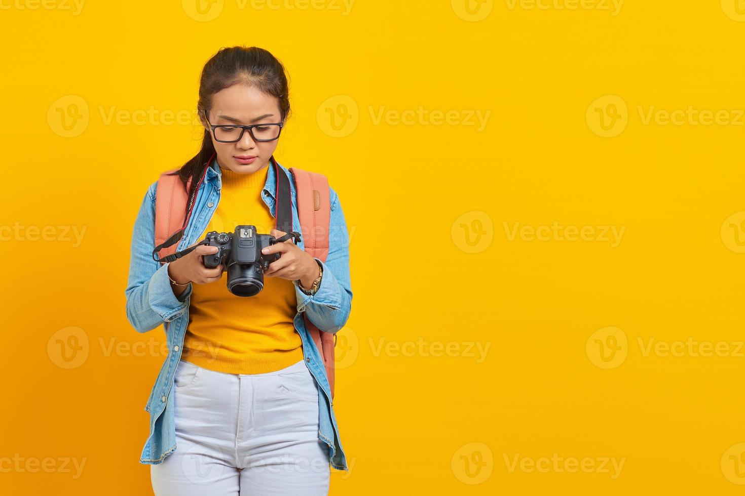 retrato de una joven asiática pensativa vestida de denim y mirando una foto en una cámara aislada de fondo amarillo. pasajero que viaja los fines de semana. concepto de viaje de vuelo aéreo