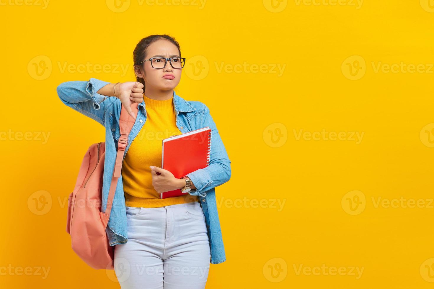 Sad young Asian woman student in denim clothes with backpack holding notebook, showing thumb down while looking aside isolated on yellow background. Education in high school university college concept photo