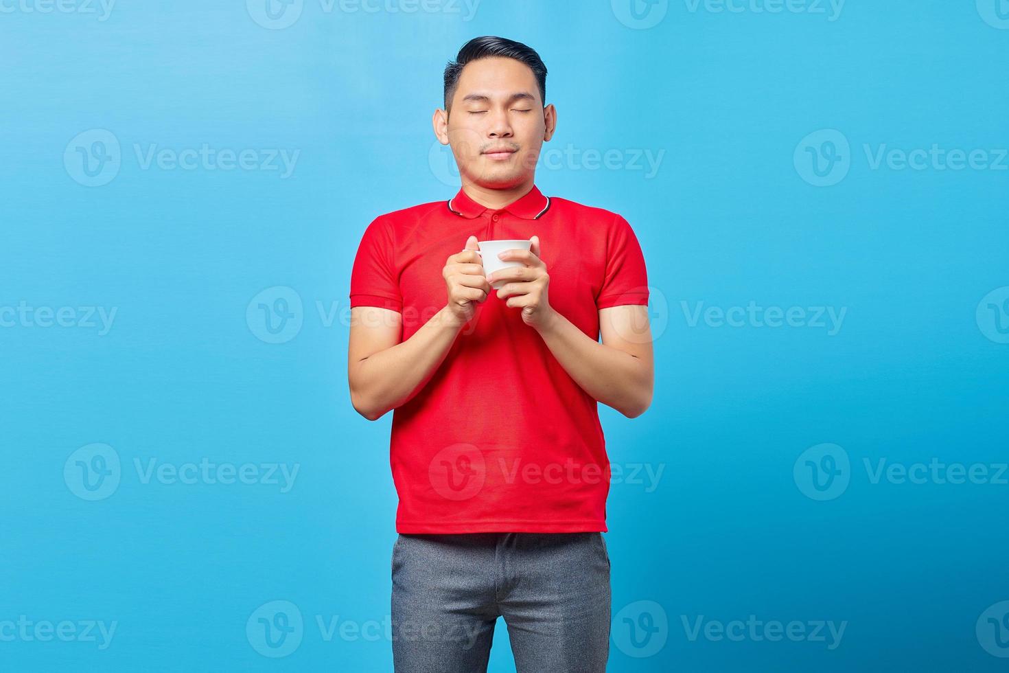 retrato de un apuesto joven asiático sosteniendo una taza de café y cerrando los ojos mientras inhala un delicioso aroma de café aislado en un fondo azul foto