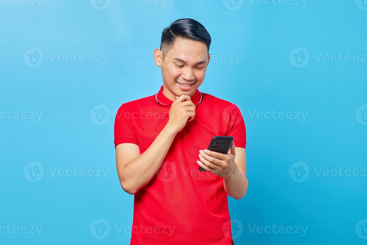 Portrait of smiling Asian young man looking at mobile phone and holding chin isolated on blue background photo
